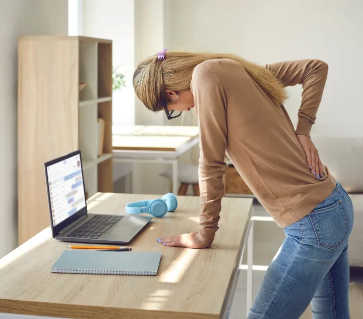 A woman leaning over a desk, holding her lower back in pain